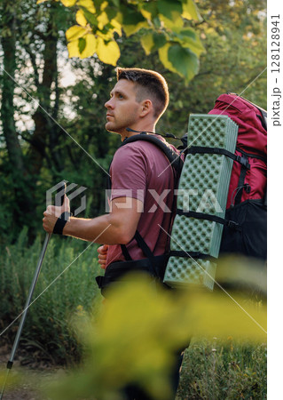 Hiker man with backpack and trekking pole walks through forest trail in sunset light. 128128941