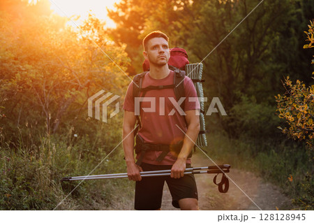 Handsome man hiker with backpack and trekking poles walking through forest at sunset. Outdoor adventure concept. 128128945