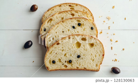 Four slices of olive-studded artisan bread lie on a white wooden surface surrounded by scattered crumbs 128130063