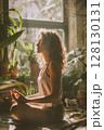 A peaceful moment as a woman practices mindfulness in a sunlit corner of her home filled with lush indoor plants. 128130131