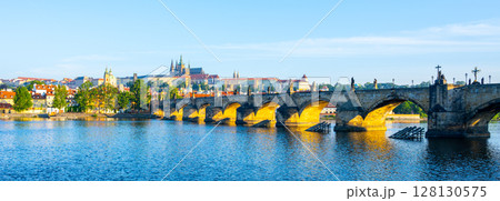 A breathtaking panorama captures the beauty of Prague Castle standing majestically above the Vltava River. Charles Bridge, illuminated by early sunlight, connects the two banks in stunning detail. 128130575