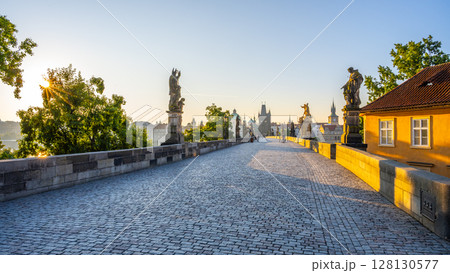 Sunlight illuminates Charles Bridge in Prague as the day begins, casting soft shadows on the cobblestones. The statues stand still, overlooking the peaceful Vltava River and vibrant embankment. 128130577