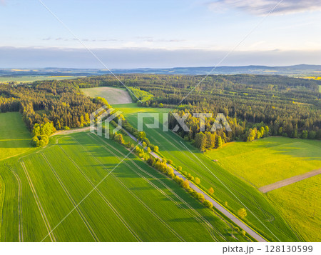 Lush green fields and forests stretch across the landscape as the sun sets on a spring evening, casting warm light over the vibrant scenery. Gentle hills create a serene backdrop. 128130599