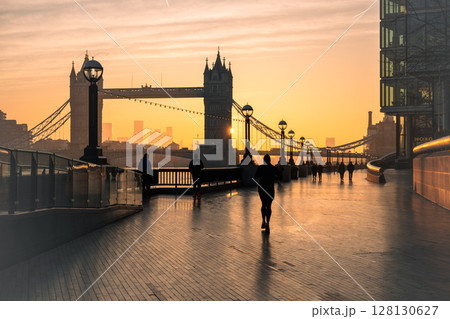 Early morning reveals Tower Bridge bathed in golden light from the rising sun. Strollers enjoy the serene atmosphere along The Queen's Walk, capturing a peaceful moment in London. 128130627