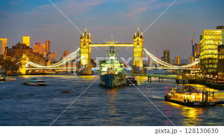 Tower Bridge illuminates in the evening sky, while HMS Belfast rests on the Thames River. Surrounding buildings glow, showcasing London's skyline after sunset. Boats navigate the serene waters. 128130630