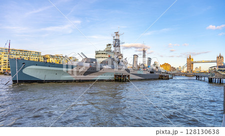HMS Belfast, a historic warship, is docked in the River Thames. The ship is surrounded by city views, including the iconic Tower Bridge on a sunny afternoon. 128130638