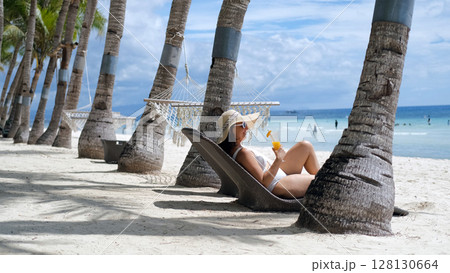 Young woman lounges on sunbed sipping mango shake by ocean on tropical beach 128130664