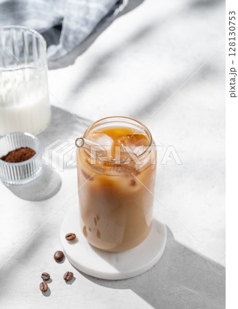 Ice latte coffee in a glass with milk on a light background with coffee beans and morning shadows. Ice latte coffee in a glass with milk on a light background with coffee beans and morning shadows. 128130753