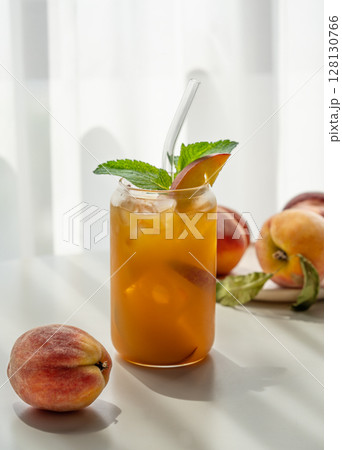 Ice tea with peach, mint in a glass on a white table near window with fresh fruits and shadow. 128130766