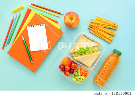 Lunchbox features sandwiches, fresh vegetables, fruits, along with stationery items like notebooks, pencils and rulers on bright blue surface, preparing for school day. 128130901