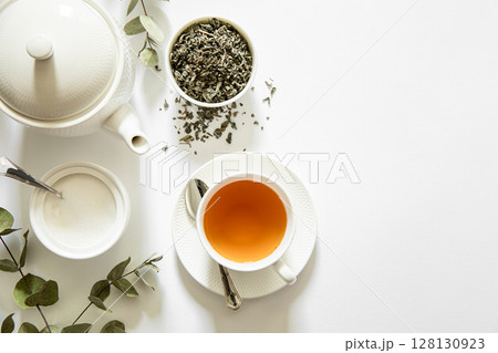 Gentle steam rises from freshly brewed tea in white cup beside herbal leaves in bowl and sugar in dish, surrounded by eucalyptus branches on bright white background. Gentle steam rises from freshly brewed tea in white cup beside herbal leaves in bowl and sugar in dish, surrounded by eucalyptus branches on bright white background. 128130923