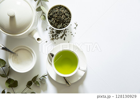 Bright green tea prepared in a simple cup alongside loose tea leaves, sugar bowl, and eucalyptus branches creating a calming atmosphere in a minimalist setting. Bright green tea prepared in a simple cup alongside loose tea leaves, sugar bowl, and eucalyptus branches creating a calming atmosphere in a minimalist setting. 128130929