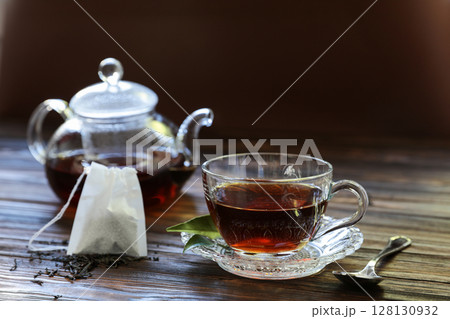 Clear glass cup filled with rich black tea sits on a saucer beside a teapot. Tea bag rests nearby, with loose tea leaves scattered on the wooden surface, inviting a moment of relaxation. 128130932