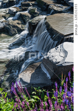 A Beautiful Tranquil Waterfall Flowing Gracefully Over Rocks Surrounded by Vibrant Flowers 128131355