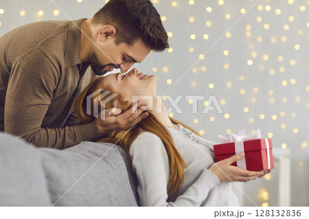 Loving man standing behind and kissing his girlfriend on the forehead, who is sitting on the couch. 128132836
