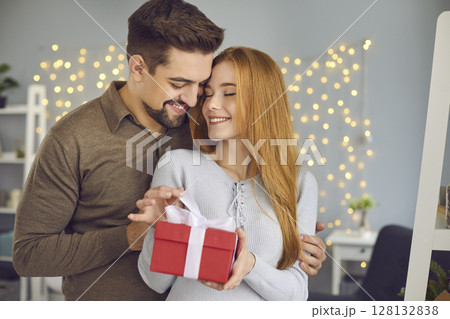 Young loving smiling couple standing, hugging and opening present box during Valentines day Young loving smiling couple standing, hugging and opening present box during Valentines day 128132838
