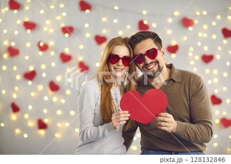 Young happy loving couple in heart shaped glasses standing and holding big red heart in hands 128132846