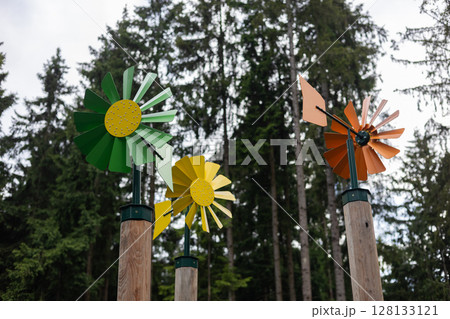 Colourful metal pinwheels spin in the wind, mounted on wooden poles against a backdrop of tall evergreen trees, creating a playful and whimsical scene in a natural setting. 128133121