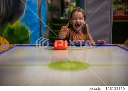 Child plays air hockey with delight. Cheerful little girl with pigtails loves to play air hockey, laughing and pointing, showing excitement and happiness in a colorful entertainment center. 128133130