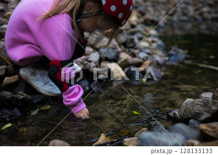 Cute child curiously explores rocky stream. Curious preschooler in helmet and protective gear touches water of shallow stream, explores nature and enjoys active recreation. 128133133