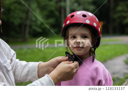 A woman fastens the straps of a red helmet on her daughter's head, protecting her before riding a bike or scooter in a green park on a summer day. Child safety while riding a bike or scooter. A woman fastens the straps of a red helmet on her daughter's head, protecting her before riding a bike or scooter in a green park on a summer day. Child safety while riding a bike or scooter. 128133137