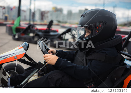 Young kart racer wearing a black helmet and protective gear, skillfully steers a go-kart around a track, focused on the race ahead and other karts parked nearby. A teenager driving a go-kart. Young kart racer wearing a black helmet and protective gear, skillfully steers a go-kart around a track, focused on the race ahead and other karts parked nearby. A teenager driving a go-kart. 128133143