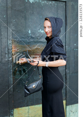 Portrait young adult elegant woman black dress and sunglasses smiles confidently in golden sunlight warm sunset backlit light against historic cathedral building. Female person walking city street 128133573