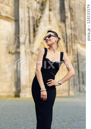 Portrait young adult elegant woman black dress and sunglasses smiles confidently in golden sunlight warm sunset backlit light against historic cathedral building. Female person walking city street 128133574