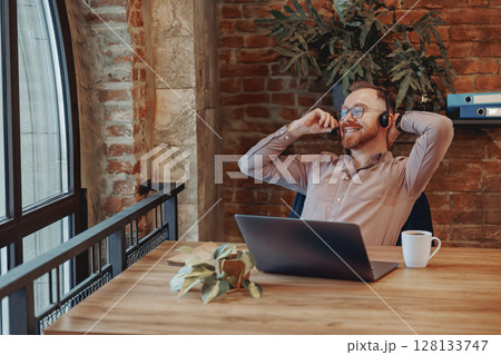 A Relaxed Professional in a Modern Workspace Utilizing a Laptop and Wearing Headphones 128133747