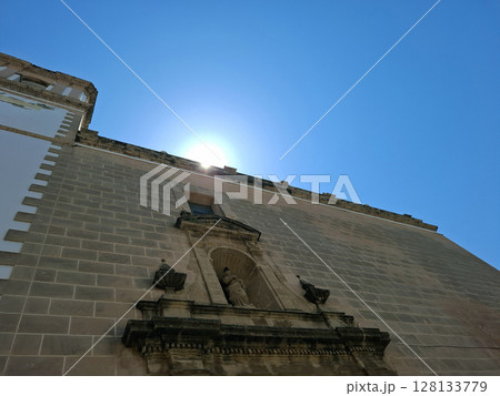 As the sun rises, golden rays light up the historical church and the statue of the saint in Denia, creating a serene morning atmosphere. Visitors can enjoy the peaceful setting. As the sun rises, golden rays light up the historical church and the statue of the saint in Denia, creating a serene morning atmosphere. Visitors can enjoy the peaceful setting. 128133779