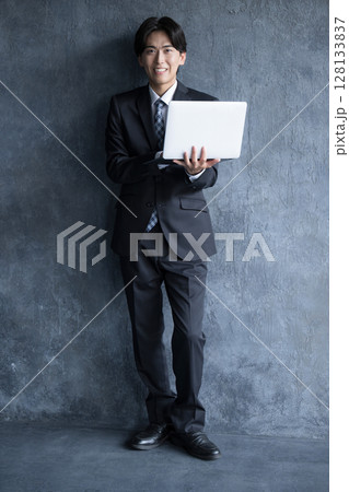 パソコンを持って考えるビジネススーツの男性　キャリアや未来,転職などを笑顔で考えるグレーバック　全身 128133837