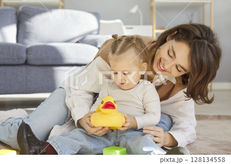 Smiling mother and little daughter playing with yellow rubber duck on floor while bonding at home. Smiling mother and little daughter playing with yellow rubber duck on floor while bonding at home. 128134558