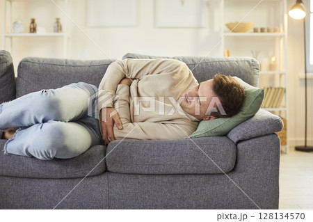 Sick young man lying on sofa, holding his stomach, suffering from abdominal pain or strong cramps. Sick young man lying on sofa, holding his stomach, suffering from abdominal pain or strong cramps. 128134570