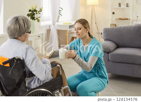 Young caregiver giving senior mature woman with disability in wheelchair a cup of beverage. Young caregiver giving senior mature woman with disability in wheelchair a cup of beverage. 128134576