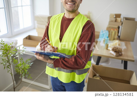Smiling male volunteer with clipboard writing notes while packing boxes and organizing donations. 128134581