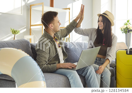 Happy couple sitting on sofa with suitcases at home booking tickets for vacation trip via laptop Happy couple sitting on sofa with suitcases at home booking tickets for vacation trip via laptop 128134584