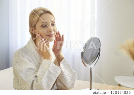 Beautiful smiling woman applying cosmetcs, cream product to face, looking in mirror, wearing robe 128134829