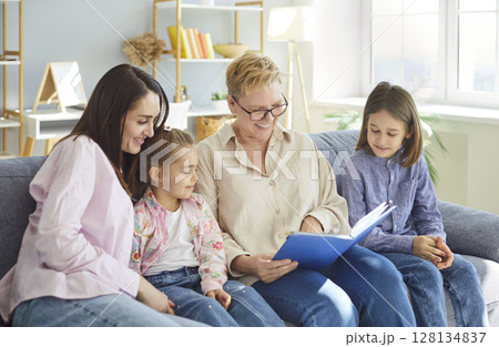 Happy family day at home, granny older woman, mother, two kids sitting on sofa reading book together Happy family day at home, granny older woman, mother, two kids sitting on sofa reading book together 128134837