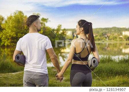 Loving fit couple, sporty people standing looking outdoors with rolled yoga mats, holding hands 128134907