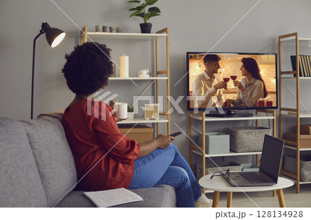 Young woman sitting on couch at home and watching romantic soap opera on television Young woman sitting on couch at home and watching romantic soap opera on television 128134928