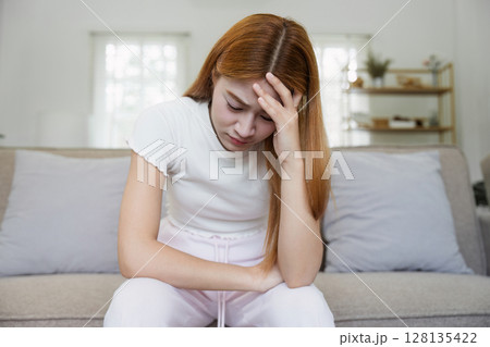 Young Woman Sitting on Couch at Home with Headache, Feeling Sick and Unwell, Holding Head in Pain Young Woman Sitting on Couch at Home with Headache, Feeling Sick and Unwell, Holding Head in Pain 128135422
