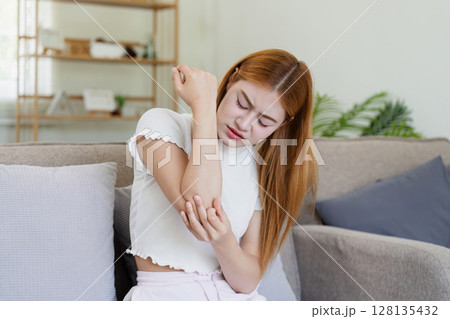 Young Woman Experiencing Elbow Pain While Sitting on a Couch at Home, Holding Her Arm in Discomfort Young Woman Experiencing Elbow Pain While Sitting on a Couch at Home, Holding Her Arm in Discomfort 128135432