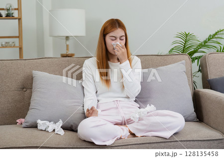 Young Woman with Cold Sitting on Couch Blowing Nose into Tissue in Cozy Home Environment Young Woman with Cold Sitting on Couch Blowing Nose into Tissue in Cozy Home Environment 128135458