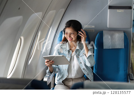 Young Woman Enjoying In-Flight Entertainment on a Modern Airplane, Smiling and Relaxed While Using a Tablet and Wearing Headphones, Capturing the Joy of Air Travel and the Comfort of Modern Aviation Young Woman Enjoying In-Flight Entertainment on a Modern Airplane, Smiling and Relaxed While Using a Tablet and Wearing Headphones, Capturing the Joy of Air Travel and the Comfort of Modern Aviation 128135577
