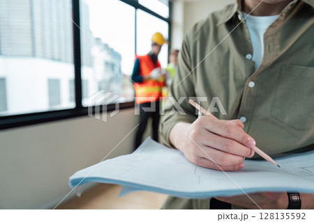 Engineer Working on Blueprint in Modern Office with Colleagues in Background, Professional Engineering Team, Construction Planning, Architectural Design, Technical Drawing, Project Management 128135592