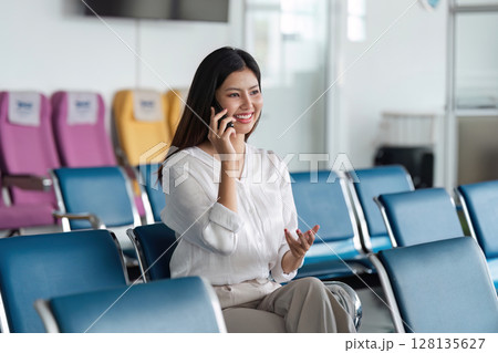 Woman Talking on Phone in Airport Lounge, Ready for Travel, Airplane Seats in Background, Modern Airport Interior, Business Travel, Waiting Area, Travel Preparation, Smiling Traveler Woman Talking on Phone in Airport Lounge, Ready for Travel, Airplane Seats in Background, Modern Airport Interior, Business Travel, Waiting Area, Travel Preparation, Smiling Traveler 128135627