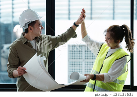 Two Engineers Collaborating on a Project, Reviewing blueprint and Celebrating Success in a Modern Office Setting with Natural Light 128135633