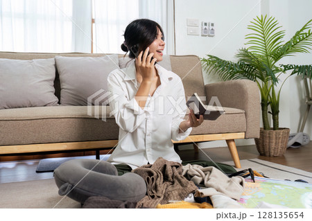 Young Woman Packing for Travel While Talking on Phone, Surrounded by Clothes and Travel Essentials in Cozy Living Room 128135654