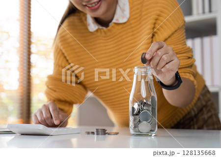 Woman Saving Money in a Jar While Calculating Bills at Home, Financial Planning and Budgeting Concept, Close-up of Hand Putting Coin in Glass Jar, Home Office Background Woman Saving Money in a Jar While Calculating Bills at Home, Financial Planning and Budgeting Concept, Close-up of Hand Putting Coin in Glass Jar, Home Office Background 128135668