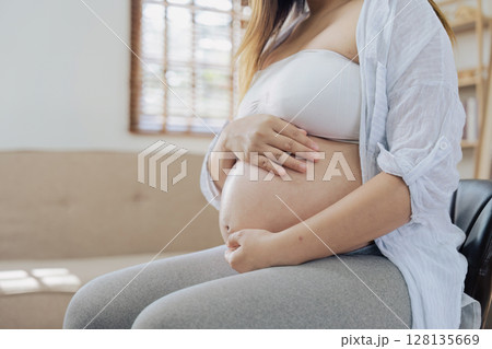 pregnancy Woman Sitting on Sofa Holding Belly, Expecting Mother in Comfortable Home Setting, Maternity and Parenthood Concept, Close Up of Baby Bump, Anticipation and Preparation for New Life pregnancy Woman Sitting on Sofa Holding Belly, Expecting Mother in Comfortable Home Setting, Maternity and Parenthood Concept, Close Up of Baby Bump, Anticipation and Preparation for New Life 128135669